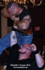 Bud Jeffries holds Pat Povilaitis aloft with one arm, while Pat bends a piece of steel.  IronMind® | Randall J. Strossen, Ph.D. photo.